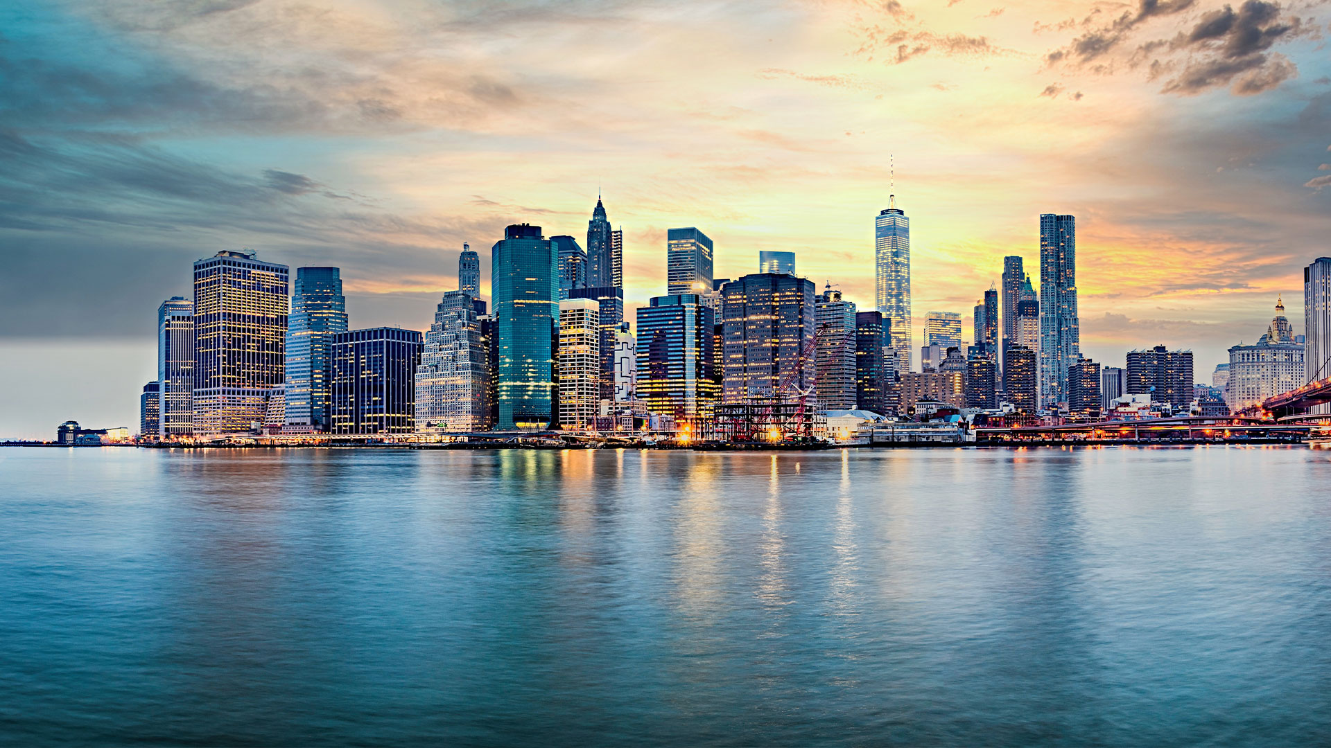 The image depicts a city skyline with the Manhattan skyline in the background and a calm river reflecting the buildings. It is a photograph taken during what appears to be either sunrise or sunset, given the warm lighting.