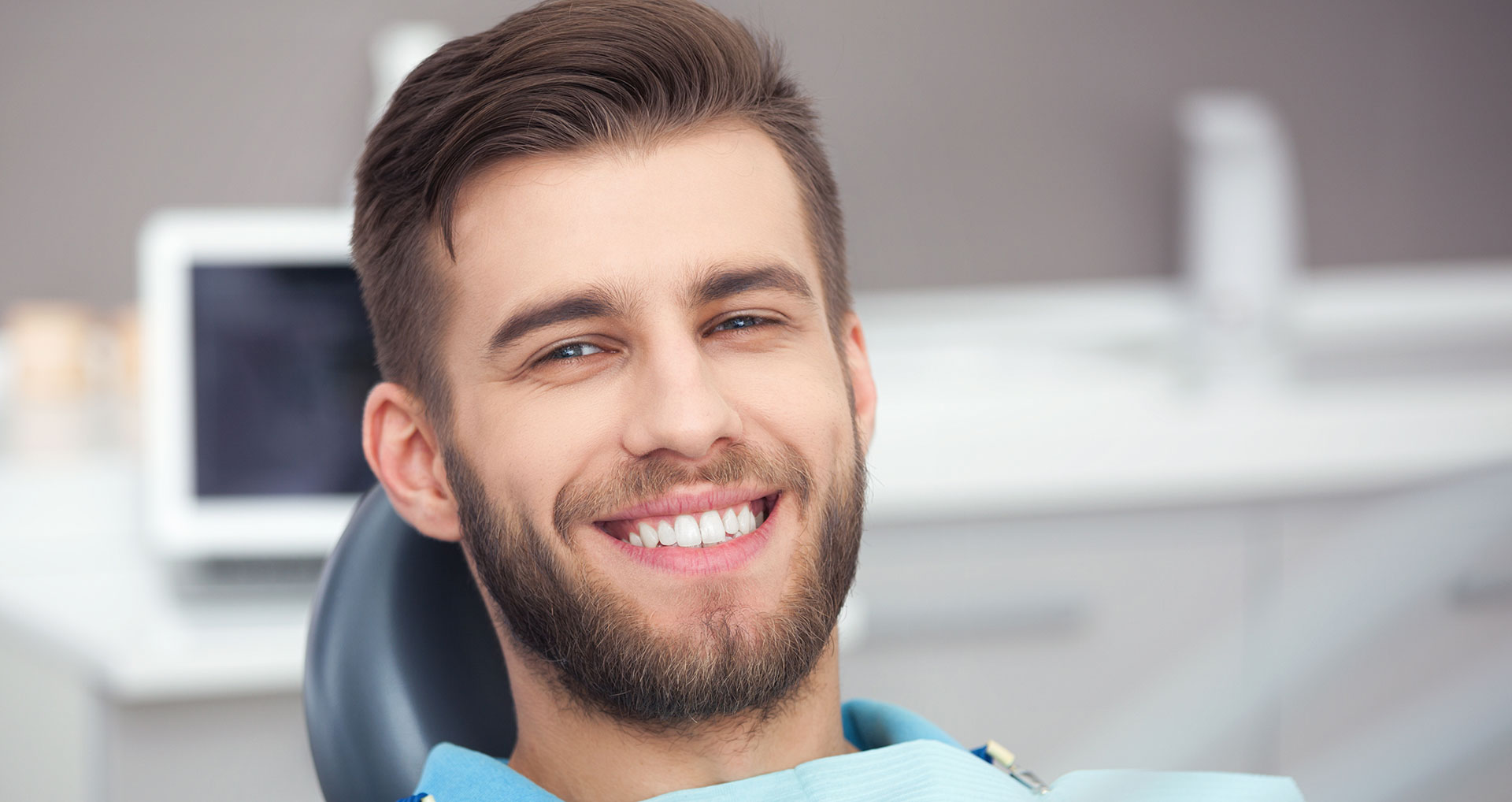 A smiling man in a dental office, wearing an apron and sitting at the chair.