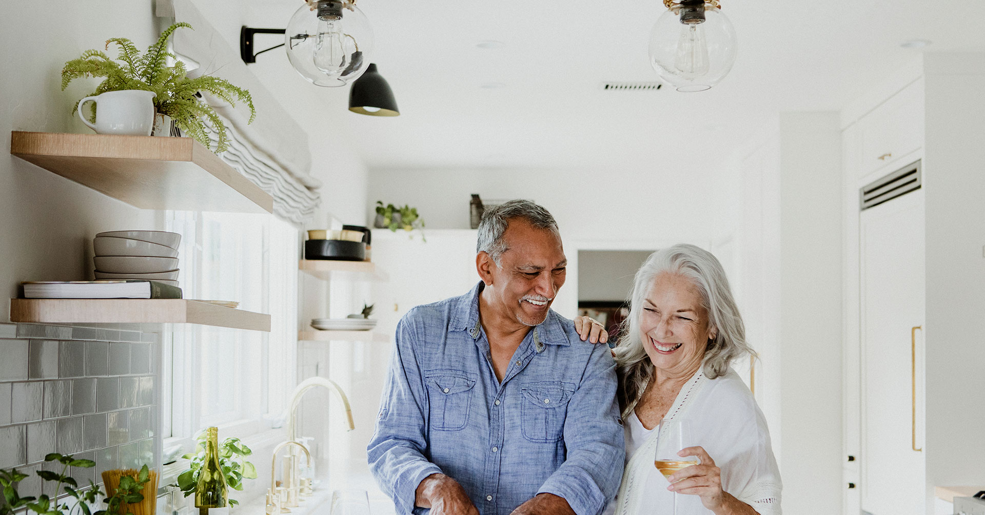 The image shows a man and woman standing in a kitchen, smiling at the camera. They are positioned near a countertop with various items on it, including a sink, a plant, and some kitchenware. The kitchen has a modern design with white cabinets, tiled walls, and a large window allowing natural light to enter.