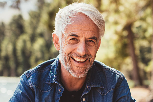 The image is a photograph of an older man with gray hair, beard, and mustache, smiling at the camera. He has a relaxed posture and appears to be outdoors, wearing a denim shirt and blue jeans.