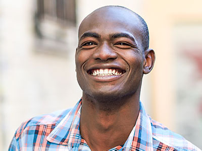 The image shows a man smiling broadly, with his eyes closed and teeth exposed, against a blurred background that suggests an outdoor setting.