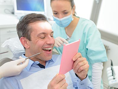 A man in a dental chair, holding a pink card and smiling while a dentist looks on.