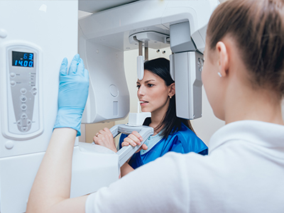 Woman in blue medical gown standing next to large white scanner, with smiling woman wearing white coat and gloves adjusting the machine.