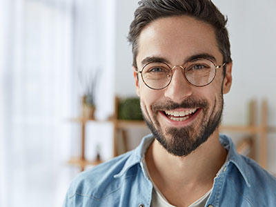 The image shows a smiling man with glasses, beard, and mustache, wearing a light blue top, standing in front of an indoor plant. He has short hair and appears to be happy or content.