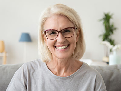 The image is a photograph of an older woman with short blonde hair, wearing glasses and a gray top. She has a smile on her face and appears to be seated indoors, possibly in a living room or a similar setting.