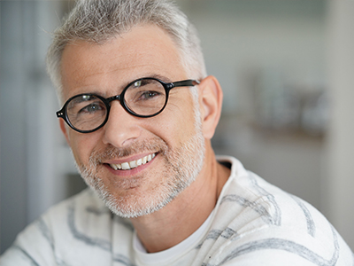 The image is a portrait of a smiling man with glasses, wearing a white top, against a blurred background that suggests an indoor setting.