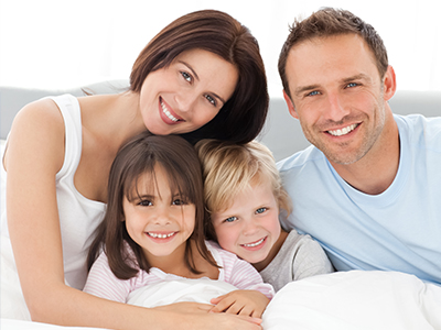 A family of five posing for a photo in bed, with the parents smiling and their children looking at the camera.