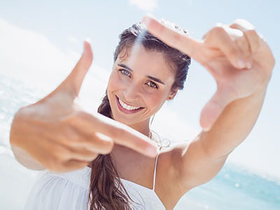 A woman is holding up a camera with both hands, smiling at the camera while looking through it.