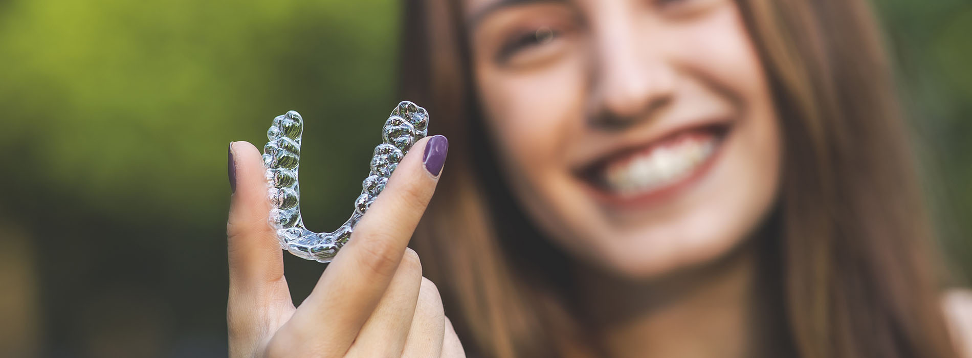 The image shows a person holding up a clear, dental-like appliance with their left hand, while smiling and looking directly at the camera.