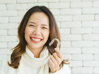 The image features a woman with a radiant smile, holding up a dental retainer with her right hand. She appears to be in a brightly lit indoor setting, possibly a dental office or a studio.